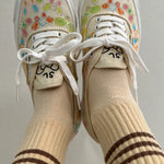 A person's fee raised in the air wearing yellow socks with brown stripes in floral sneakers with light grey background