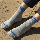 A pair of grey ankle crew socks with blue stripes, displayed on a person's feet against a textured background.