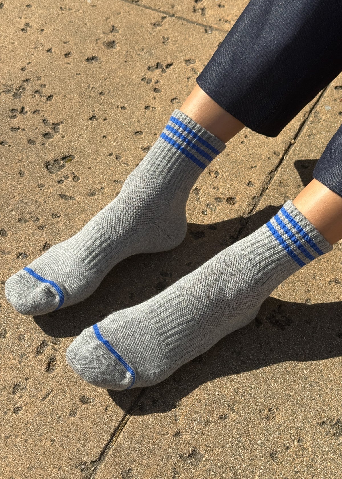 A pair of grey ankle crew socks with blue stripes, displayed on a person's feet against a textured background.