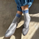 Person wearing grey socks with blue stripes, grey shoes and blue pants on concrete floor with sun shining on the feet