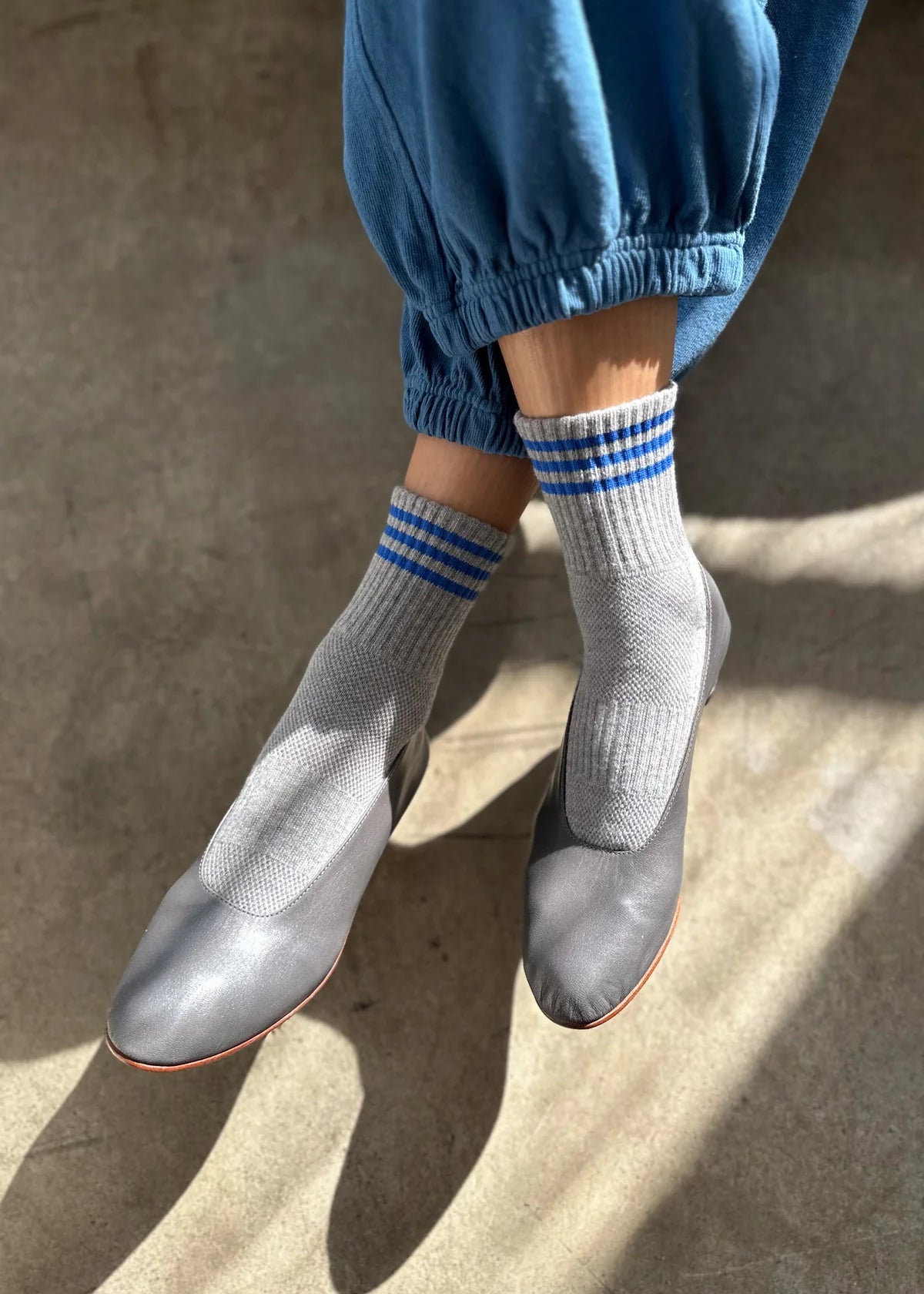 Person wearing grey socks with blue stripes, grey shoes and blue pants on concrete floor with sun shining on the feet