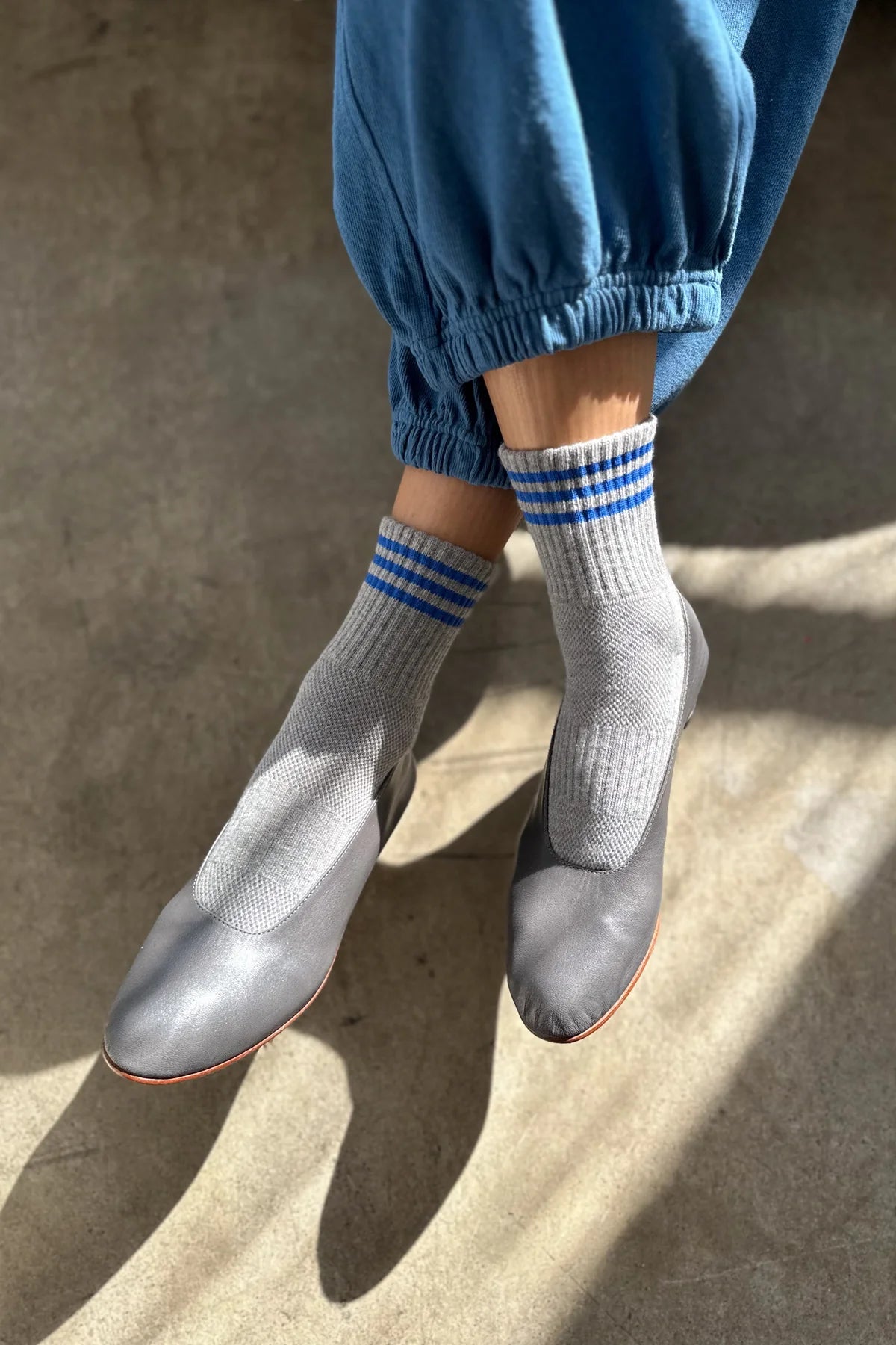 Person wearing grey socks with blue stripes, grey shoes and blue pants on concrete floor with sun shining on the feet