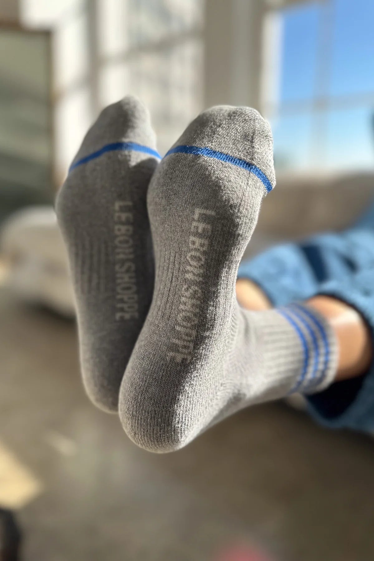 Person wearing grey socks with blue stripes and Le Bon Shoppe branding with windows in the background