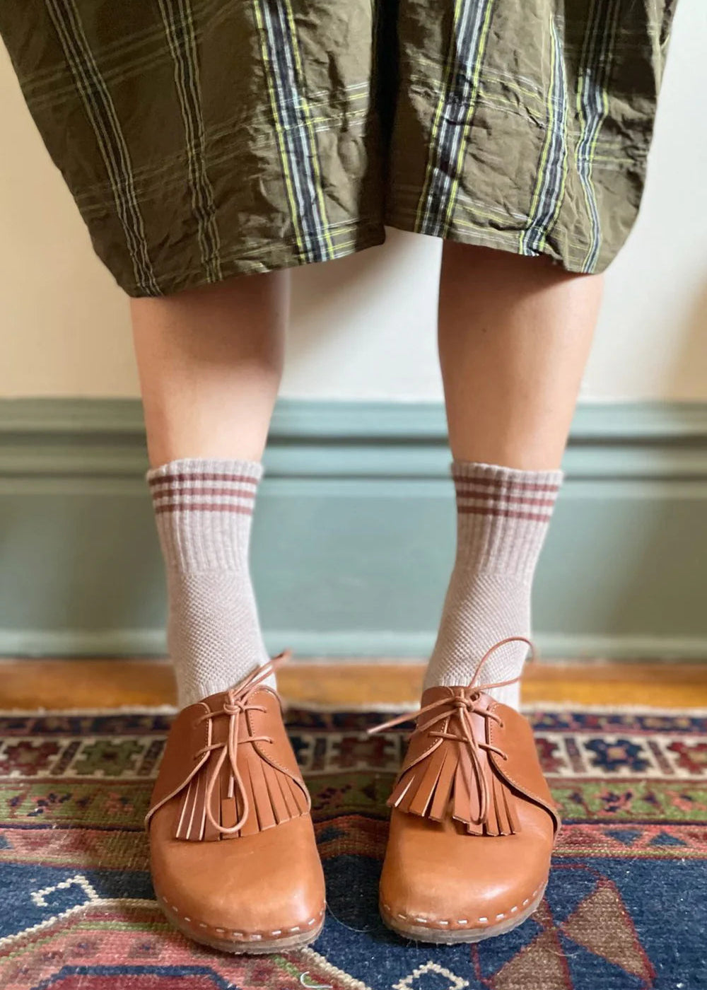 Person wearing brown shoes with fringes, plaid skirt, and checkered socks on a patterned rug.