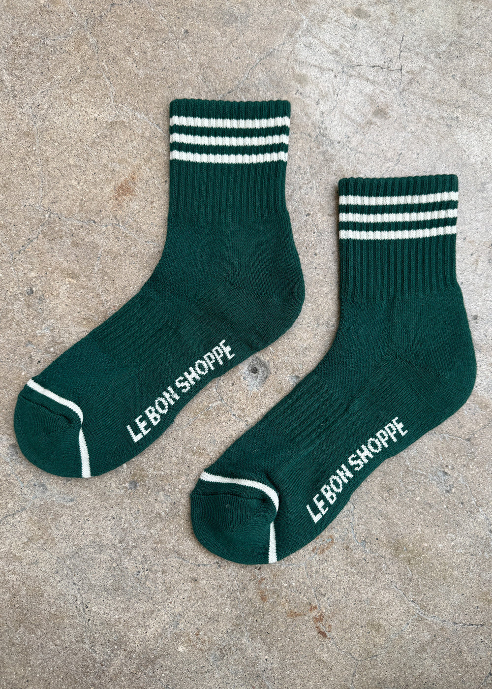 Green socks with white stripes and white Le Bon Shoppe branding on a concrete background