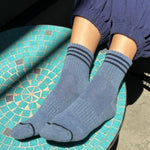 A person wearing indigo ankle crew socks with triple contrast stripes, seated on a tile surface.