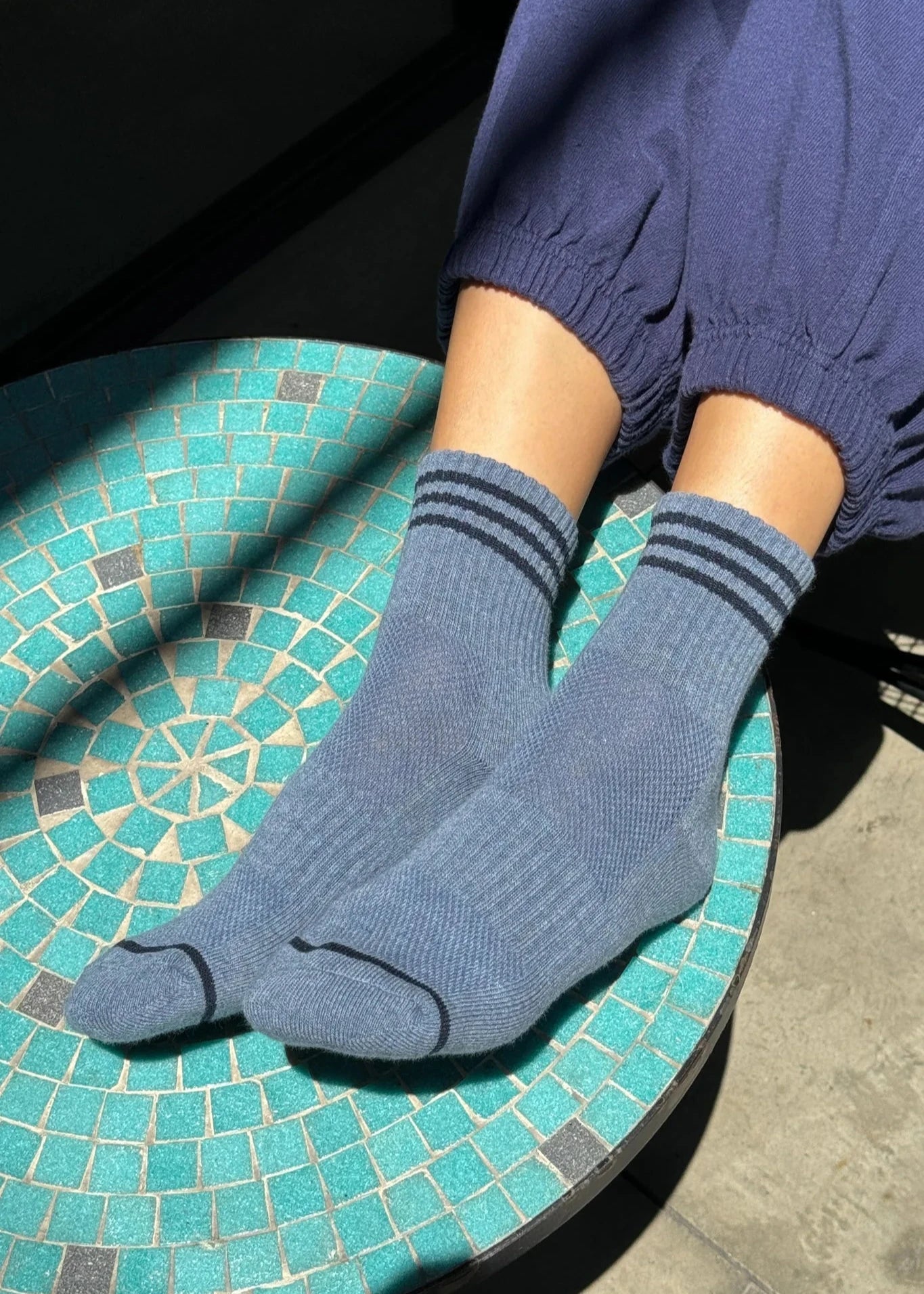 A person wearing indigo ankle crew socks with triple contrast stripes, seated on a tile surface.