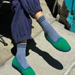 Person wearing blue pants, blue socks with navy stripes and green slip-on shoes on sidewalk
