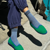 Person wearing blue pants, blue socks with navy stripes and green slip-on shoes on sidewalk