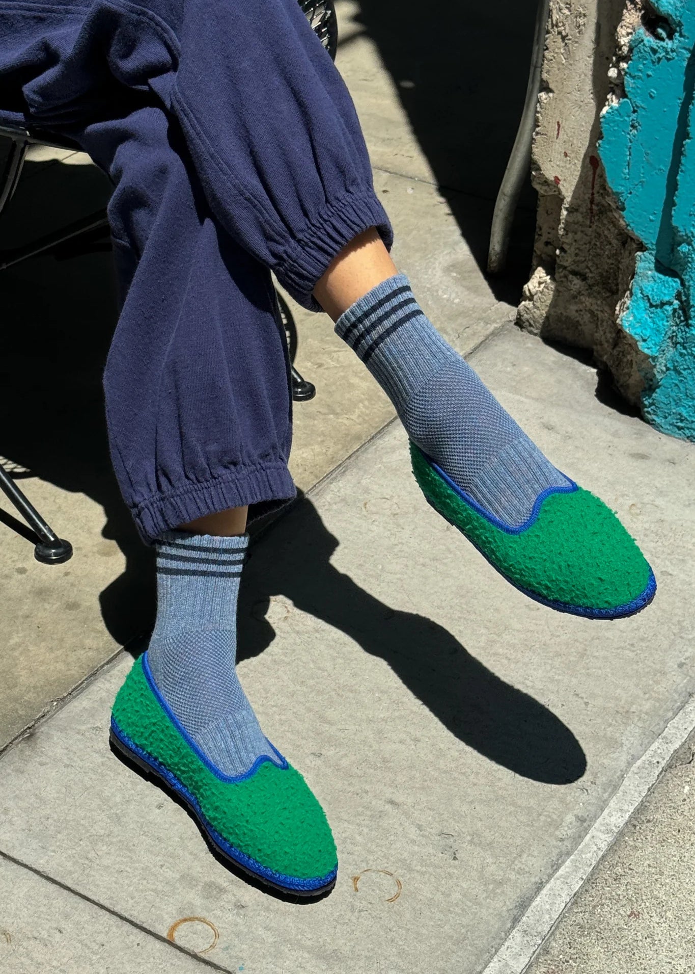 Person wearing blue pants, blue socks with navy stripes and green slip-on shoes on sidewalk