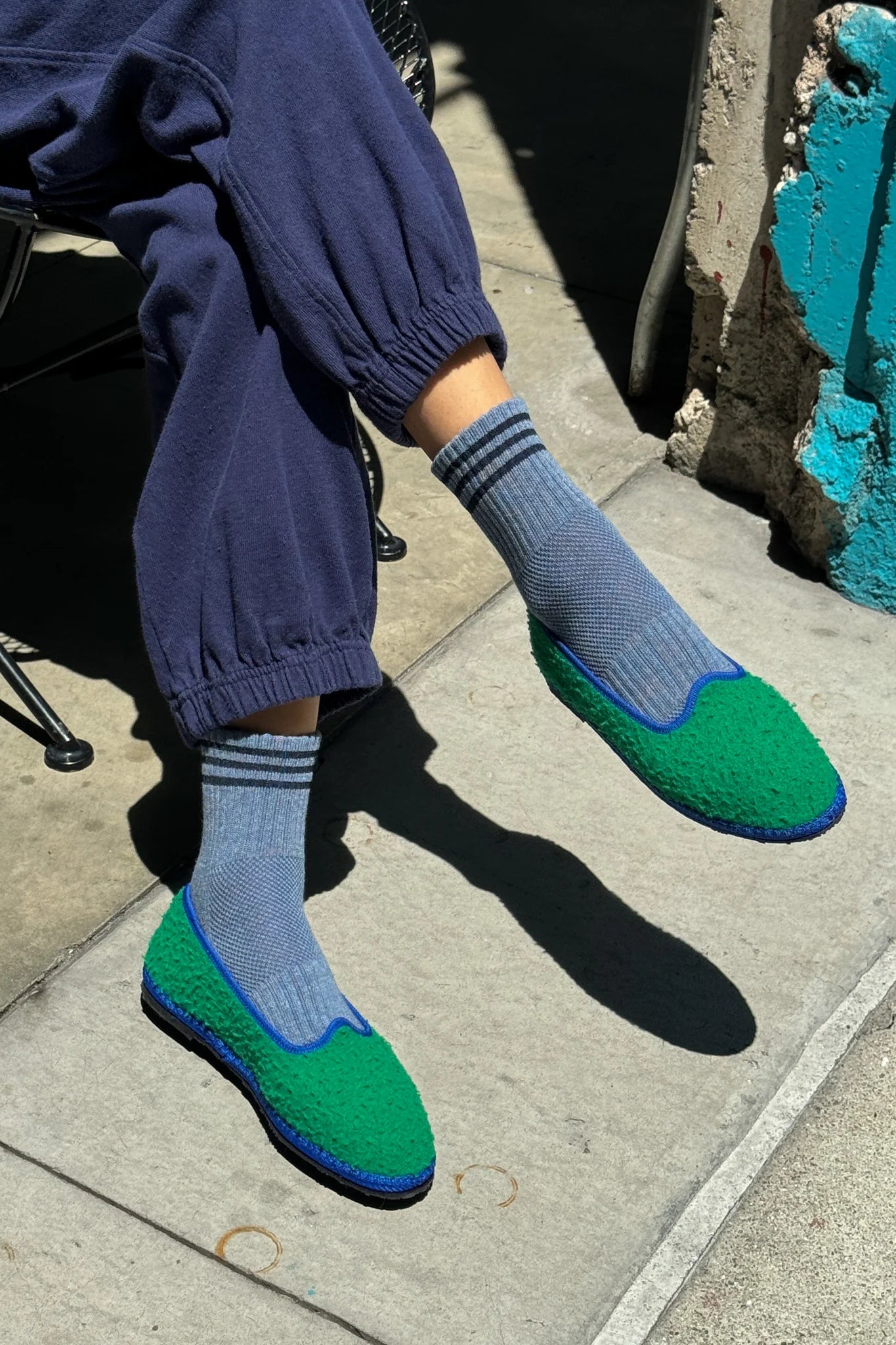 Person wearing blue pants, blue socks with navy stripes and green slip-on shoes on sidewalk