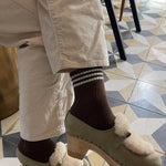 Person wearing cream colored pants, brown socks and taupe mules on tile checked floor