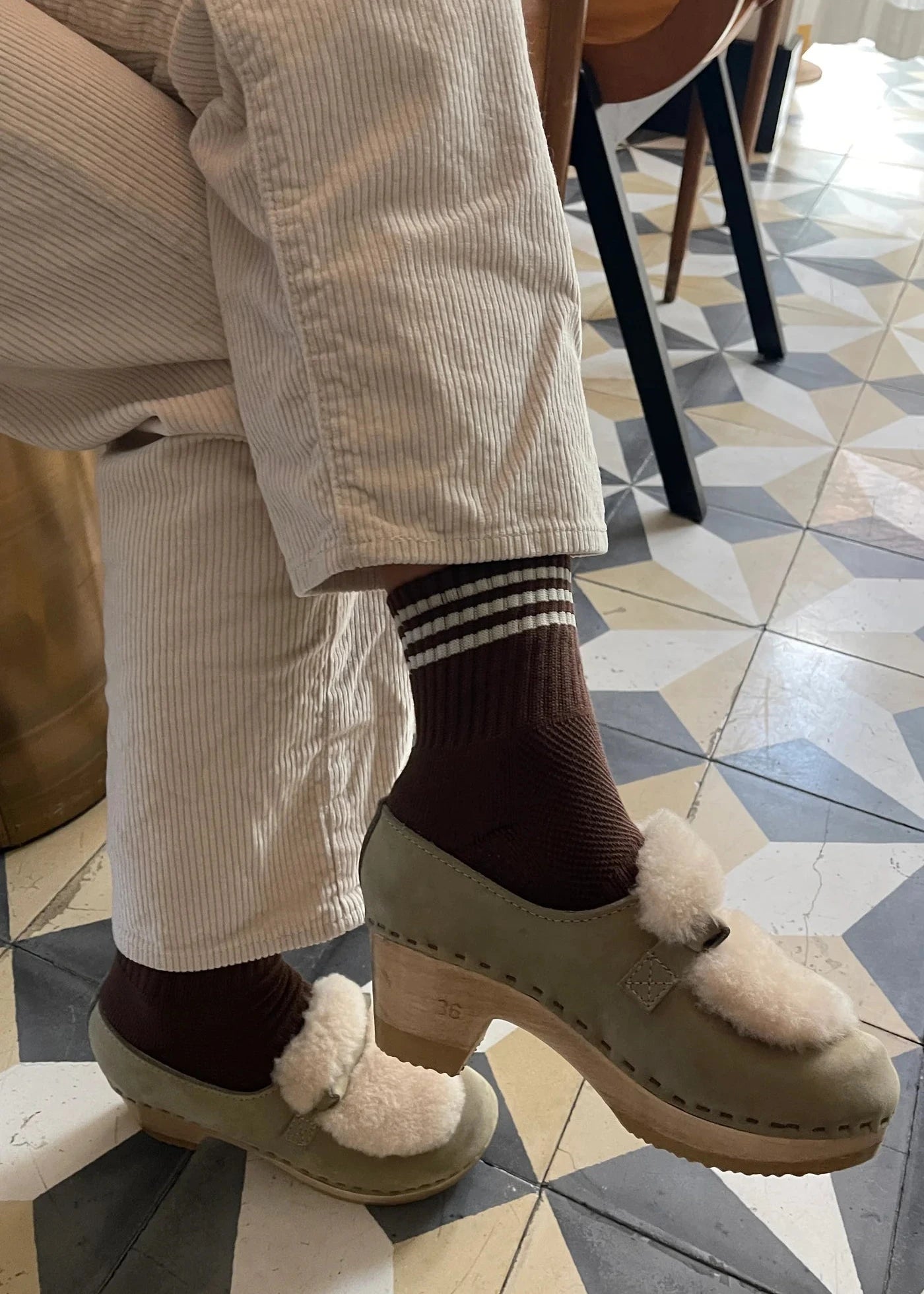 Person wearing cream colored pants, brown socks and taupe mules on tile checked floor