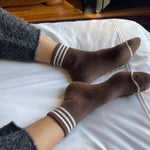 person wearing grey pants and brown socks with white stripes on a bed with white sheets