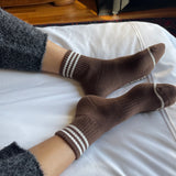 person wearing grey pants and brown socks with white stripes on a bed with white sheets