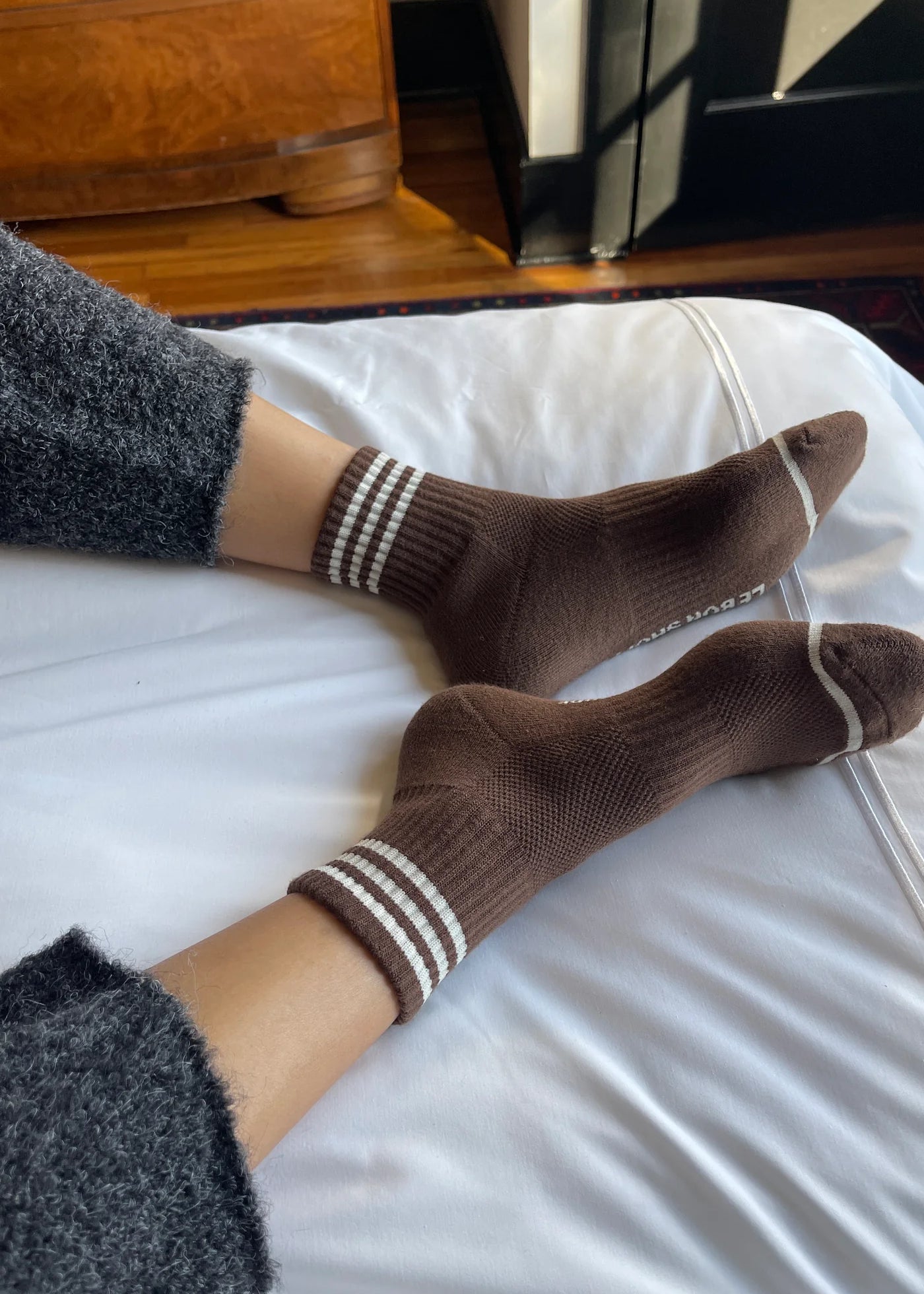 person wearing grey pants and brown socks with white stripes on a bed with white sheets