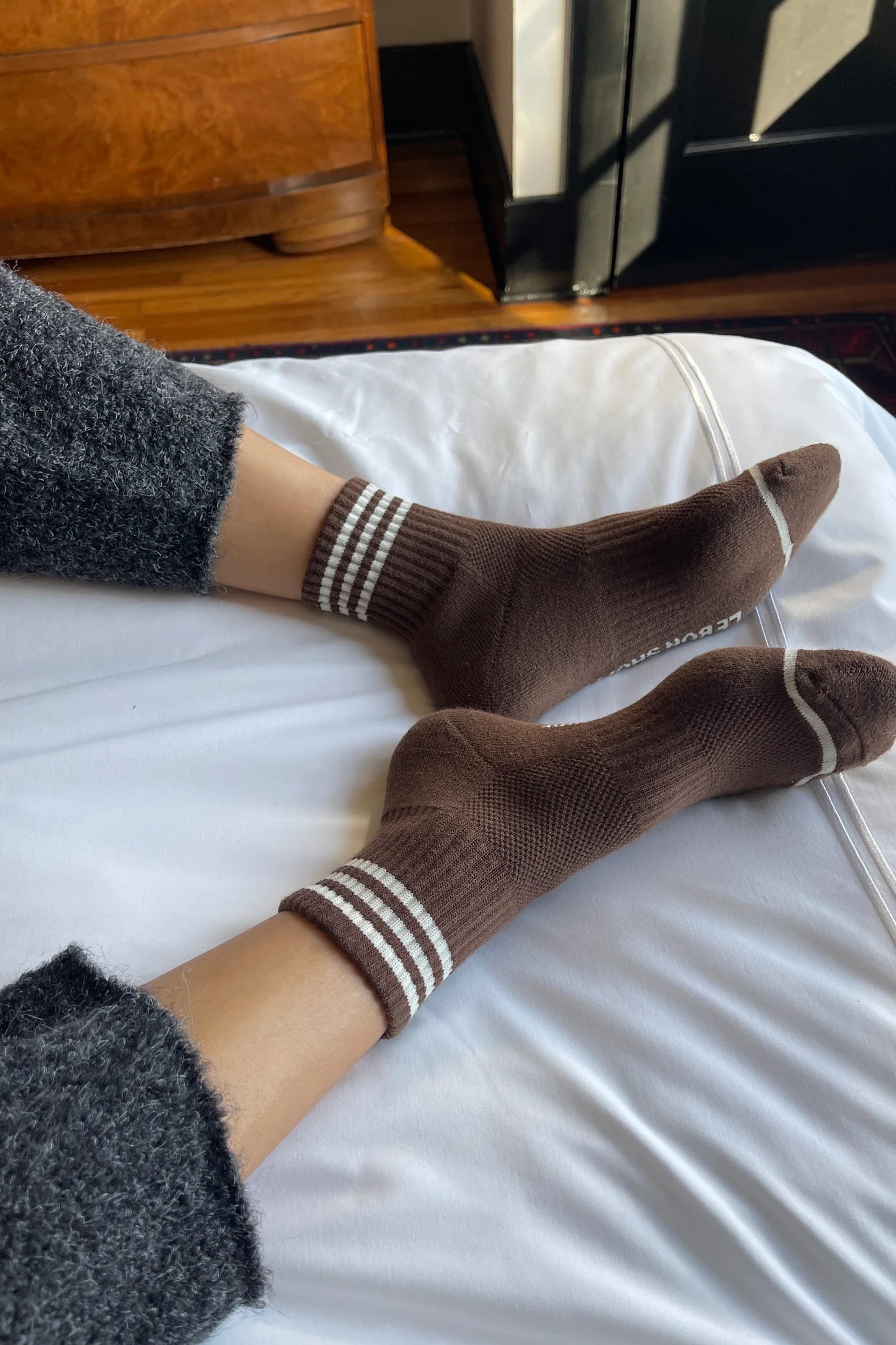 person wearing grey pants and brown socks with white stripes on a bed with white sheets