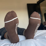 person wearing grey pants and brown socks with white stripes on a bed with white sheets