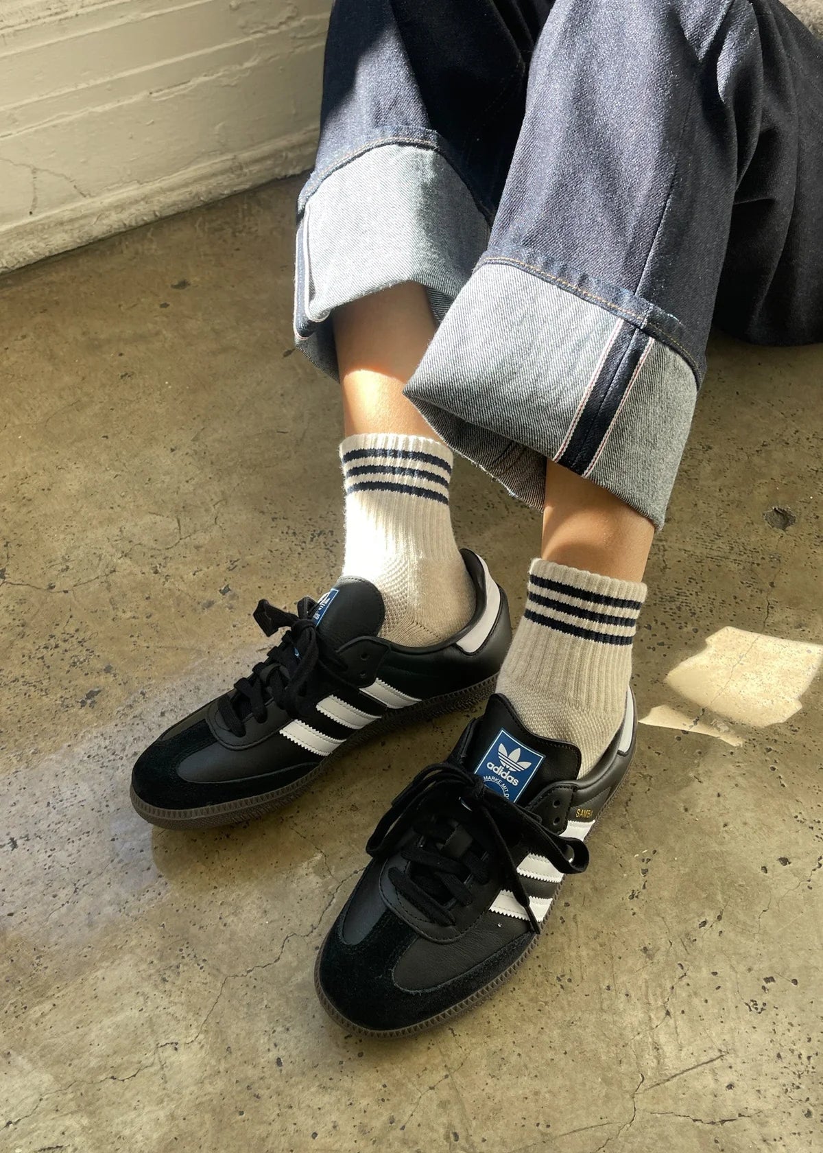 Person wearing black Adidas sneakers with white stripes on a concrete floor.