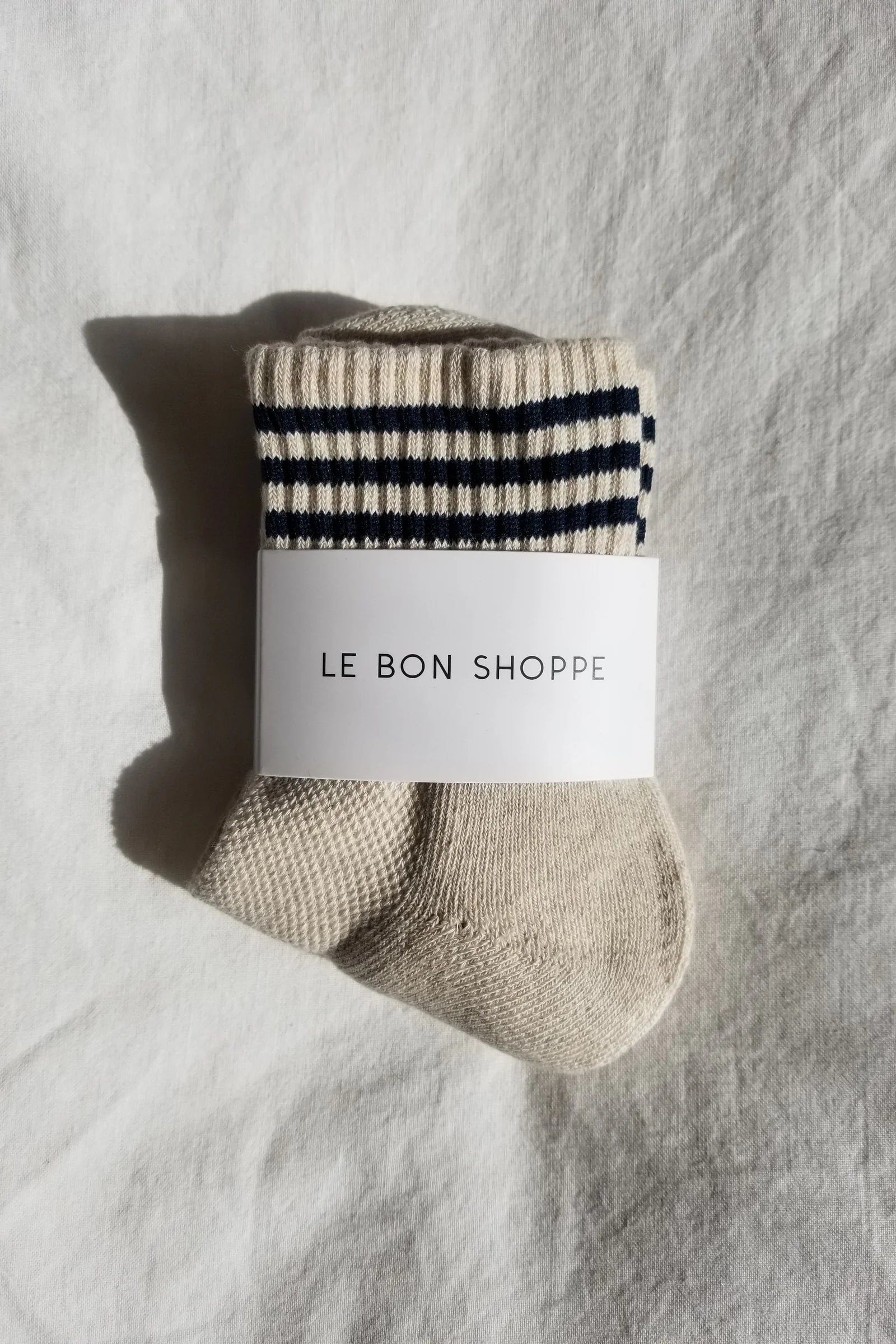 Beige sock with black stripes and 'Le Bon Shoppe' label on a textured gray background