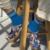 person sitting on a woodd stool wearing blue socks with white stripes and floral clogs