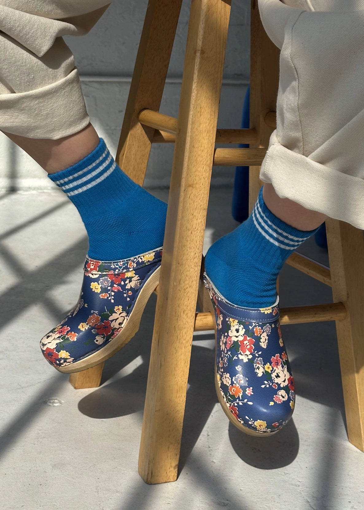 person sitting on a woodd stool wearing blue socks with white stripes and floral clogs