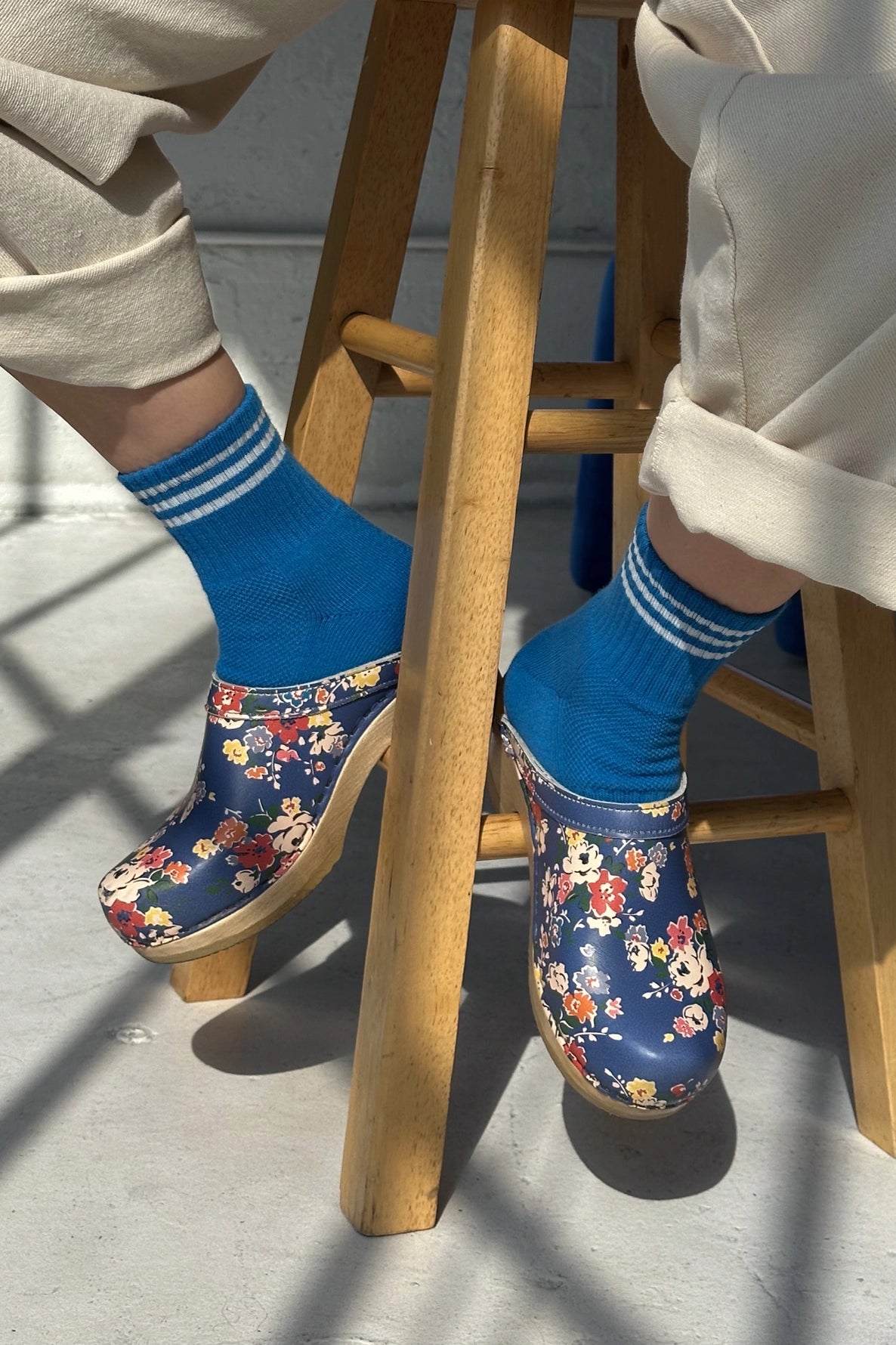 person sitting on a woodd stool wearing blue socks with white stripes and floral clogs