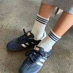 A pair of white ankle socks with a blue  stripes at the opening, worn with gray sneakers.