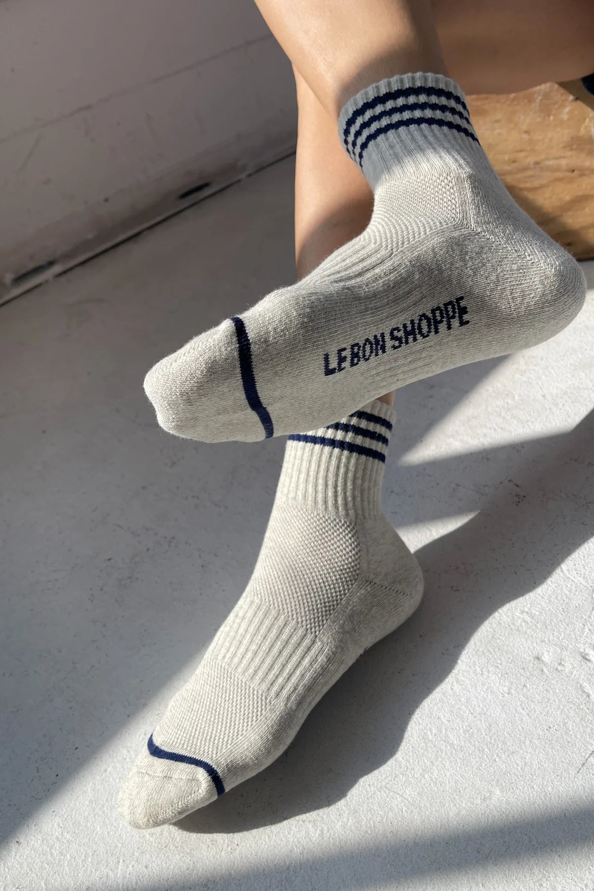 Person wearing oatmeal socks with blue stripes and Le Bon Shoppe branding on light grey background