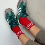A pair of red ankle crew socks with white and green stripes at the heel and toe, worn with green sneakers.