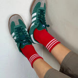 A pair of red ankle crew socks with white and green stripes at the heel and toe, worn with green sneakers.