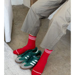 Person wearing grey pants andred socks with white stripes . There are green sneakers between their feet.