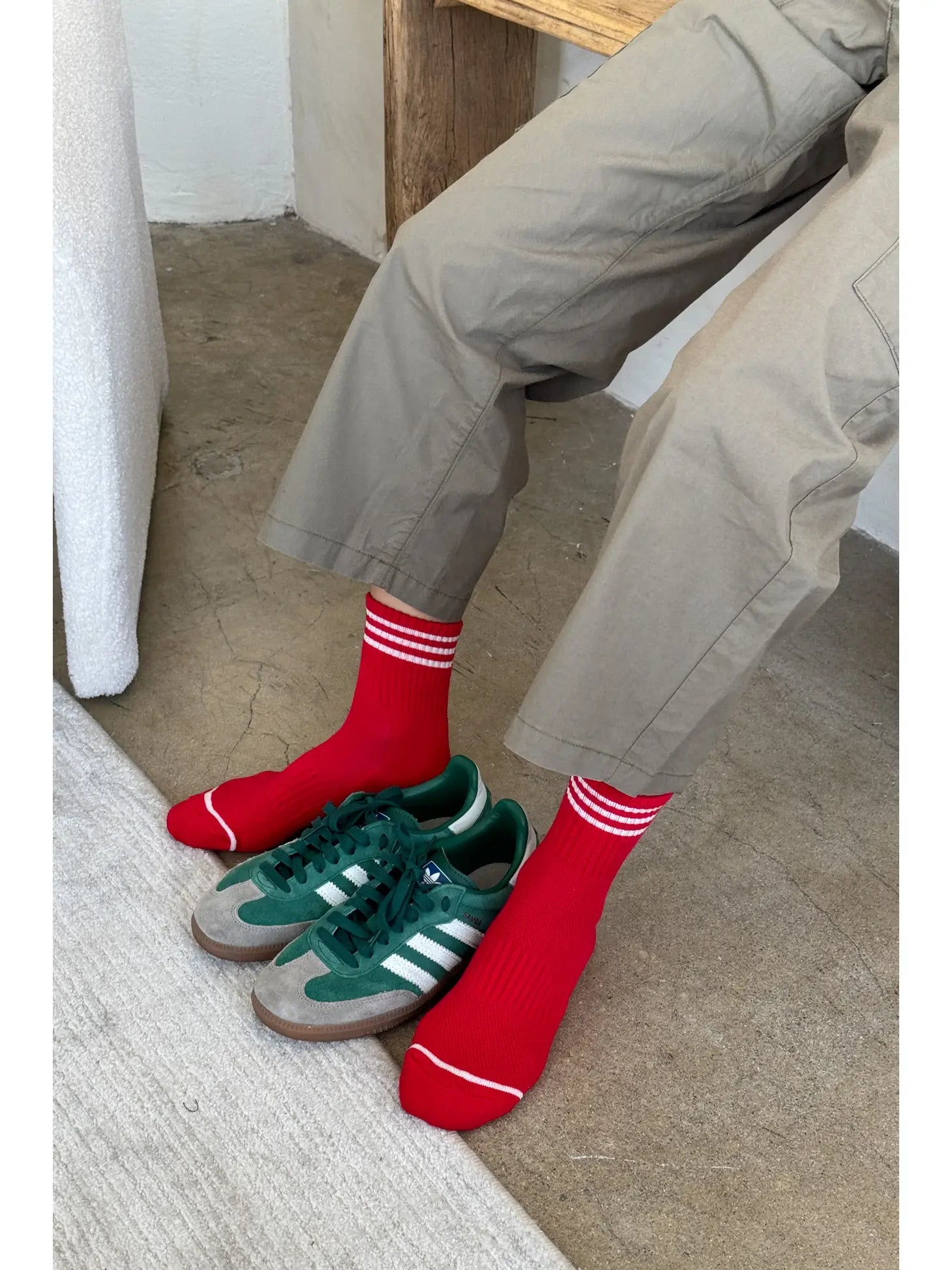 Person wearing grey pants andred socks with white stripes . There are green sneakers between their feet.