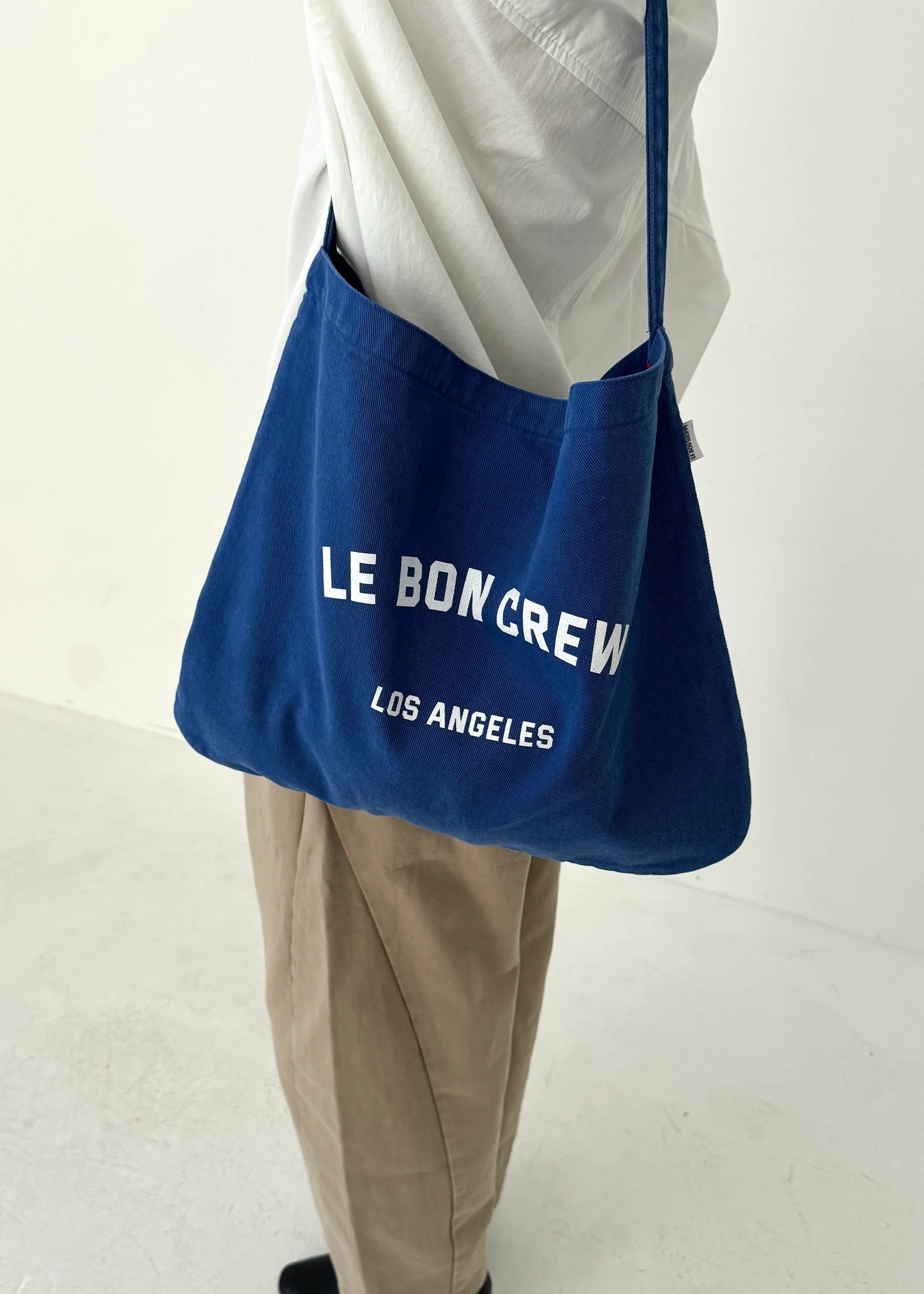 Blue bag with 'LE BON CREW' text held by a person wearing beige pants on a white background