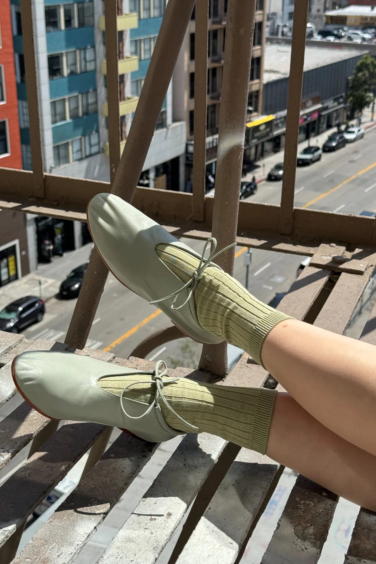 Person wearing light green shoes and socks on a balcony with a city street view.