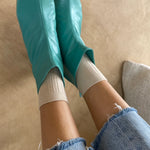Teal rubber booties worn with white socks and blue jeans on a beige floor.