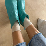 Teal rubber booties worn with white socks and blue jeans on a beige floor.