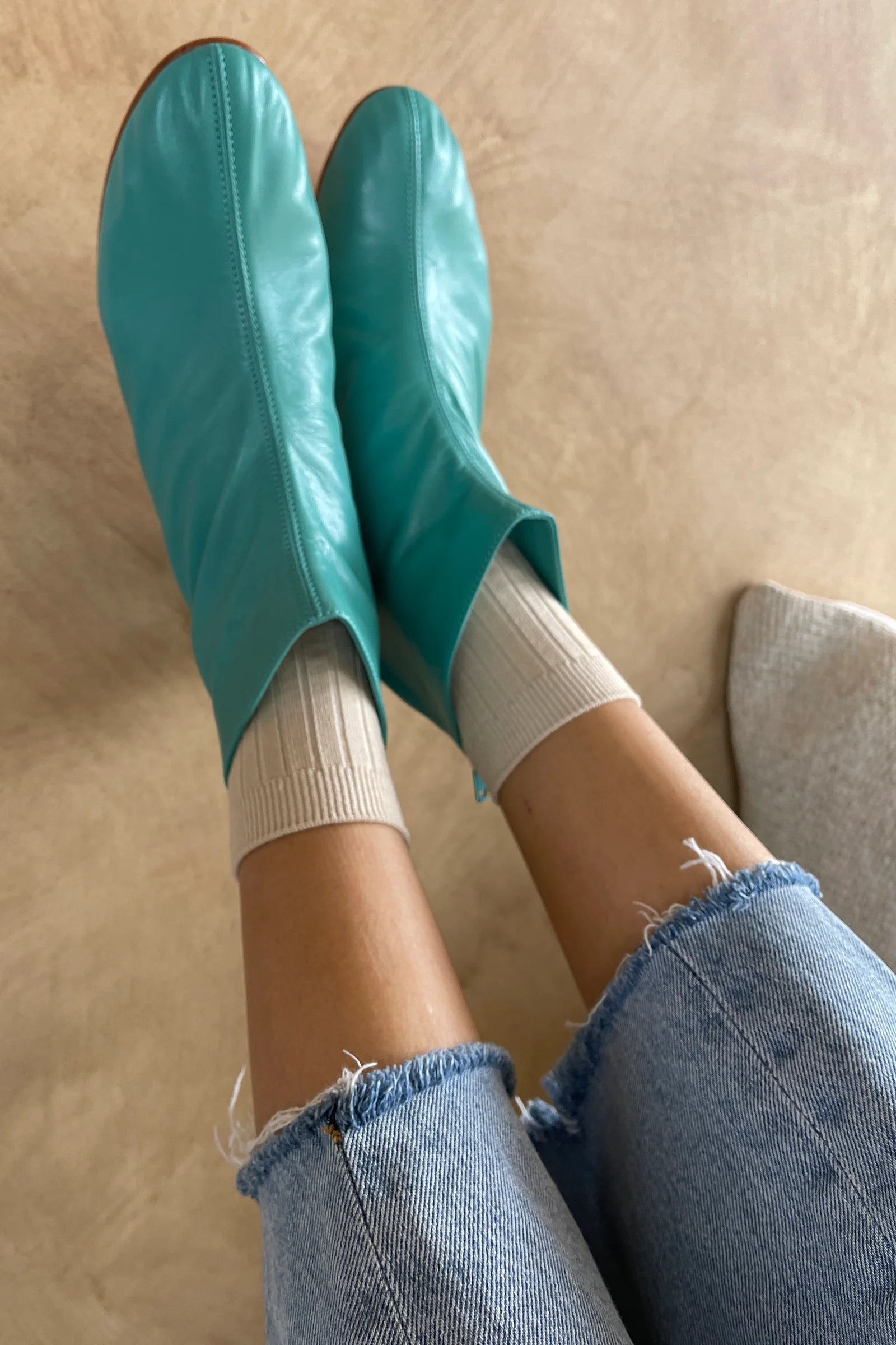 Teal rubber booties worn with white socks and blue jeans on a beige floor.