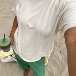 Person wearing a white t-shirt and green pants, holding a drink on a sandy surface.