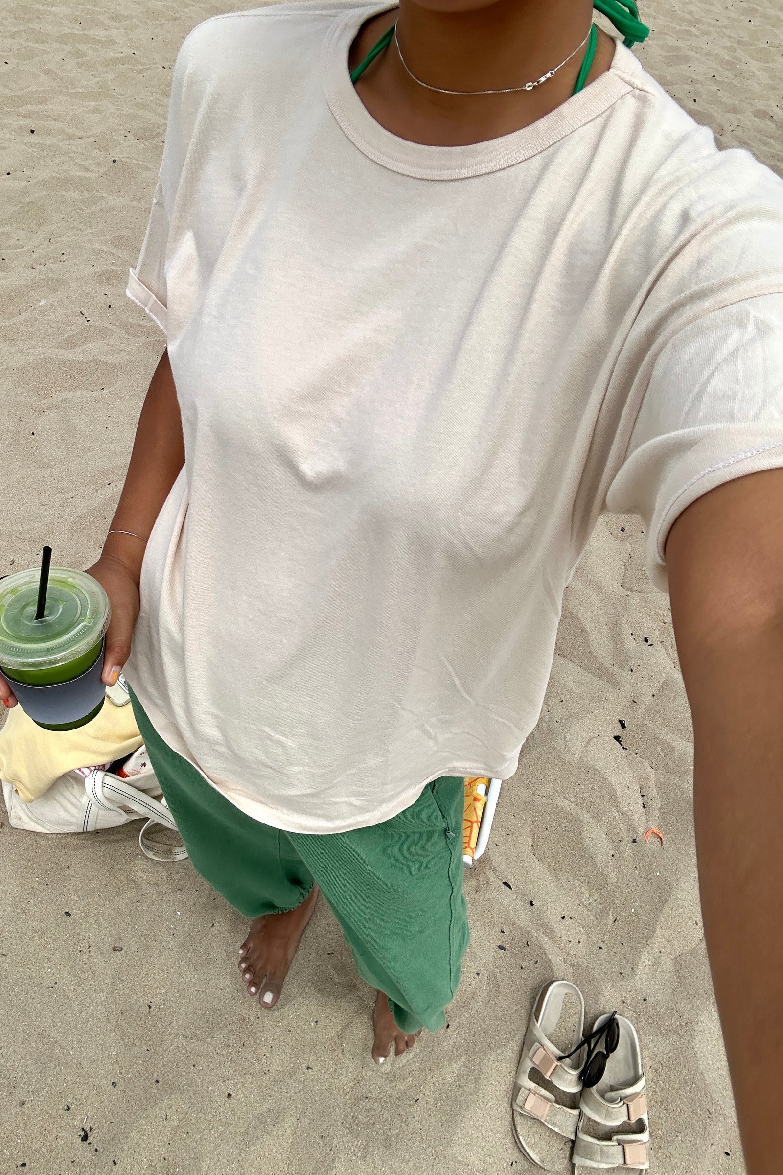 Person wearing a white t-shirt and green pants, holding a drink on a sandy surface.