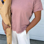 Person wearing a pink t-shirt and white pants with a beige jacket, standing against a white wall.
