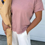 Person wearing a pink t-shirt and white pants with a beige jacket, standing against a white wall.