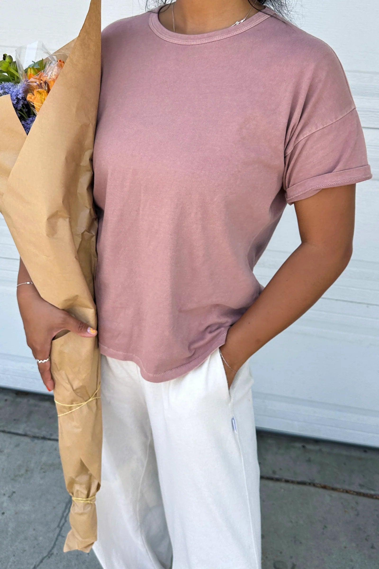Person wearing a pink t-shirt and white pants with a beige jacket, standing against a white wall.