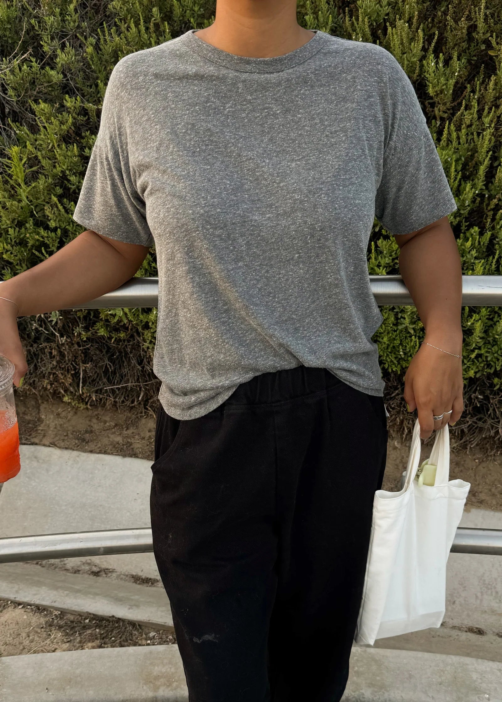 Person wearing a gray t-shirt and black pants holding a white bag outdoors.