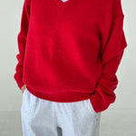 Person wearing a red sweater over a white shirt with gray pants against a plain background