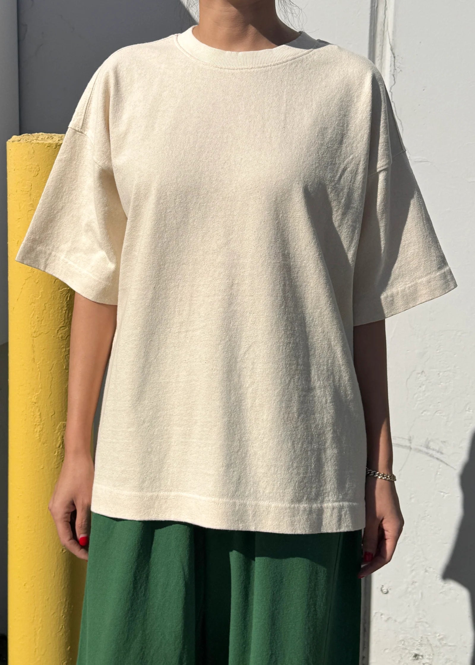 Person wearing a beige oversized t-shirt and green pants against a white wall.
