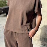 Person wearing a brown t-shirt and pants standing against a concrete wall.