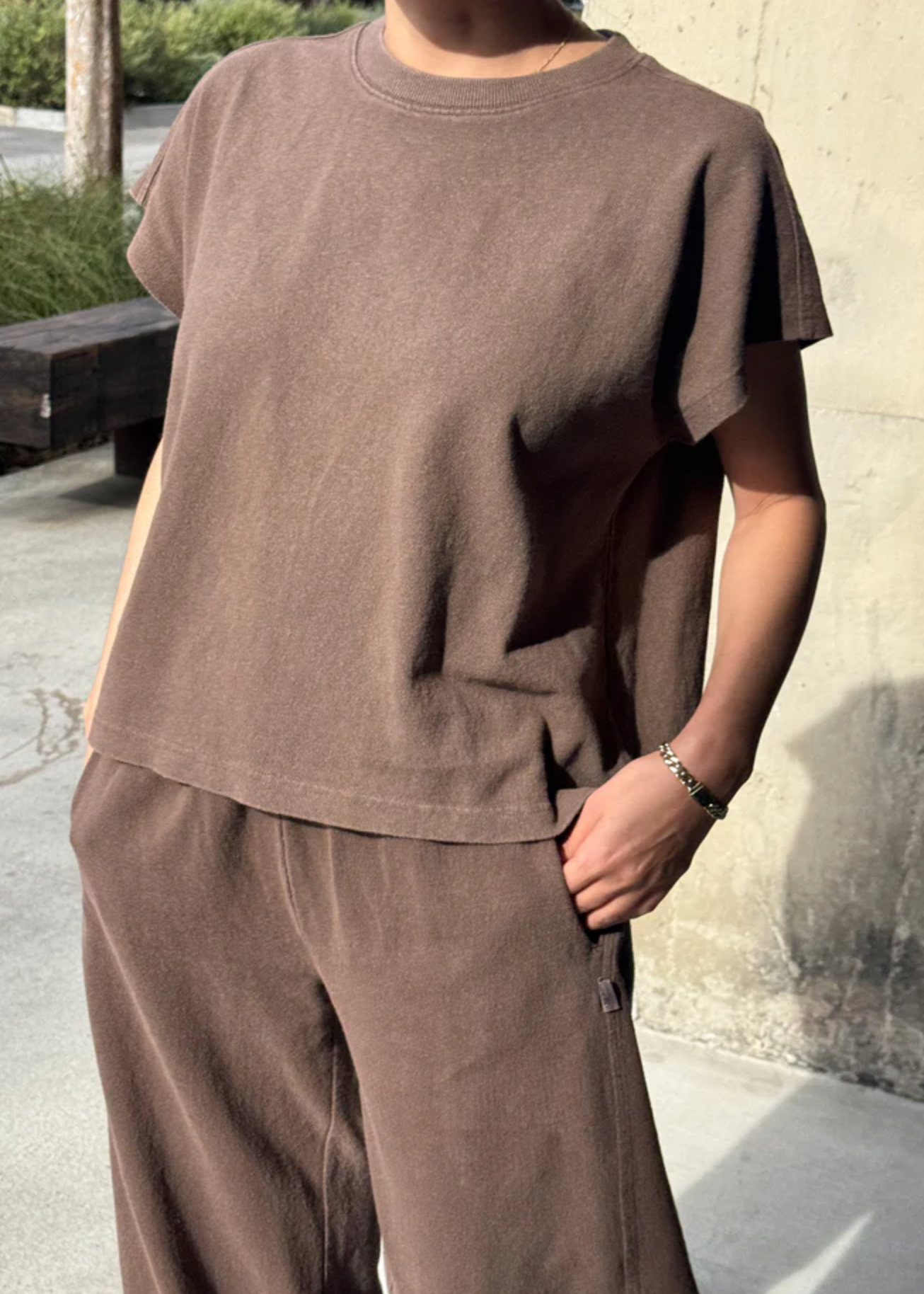 Person wearing a brown t-shirt and pants standing against a concrete wall.