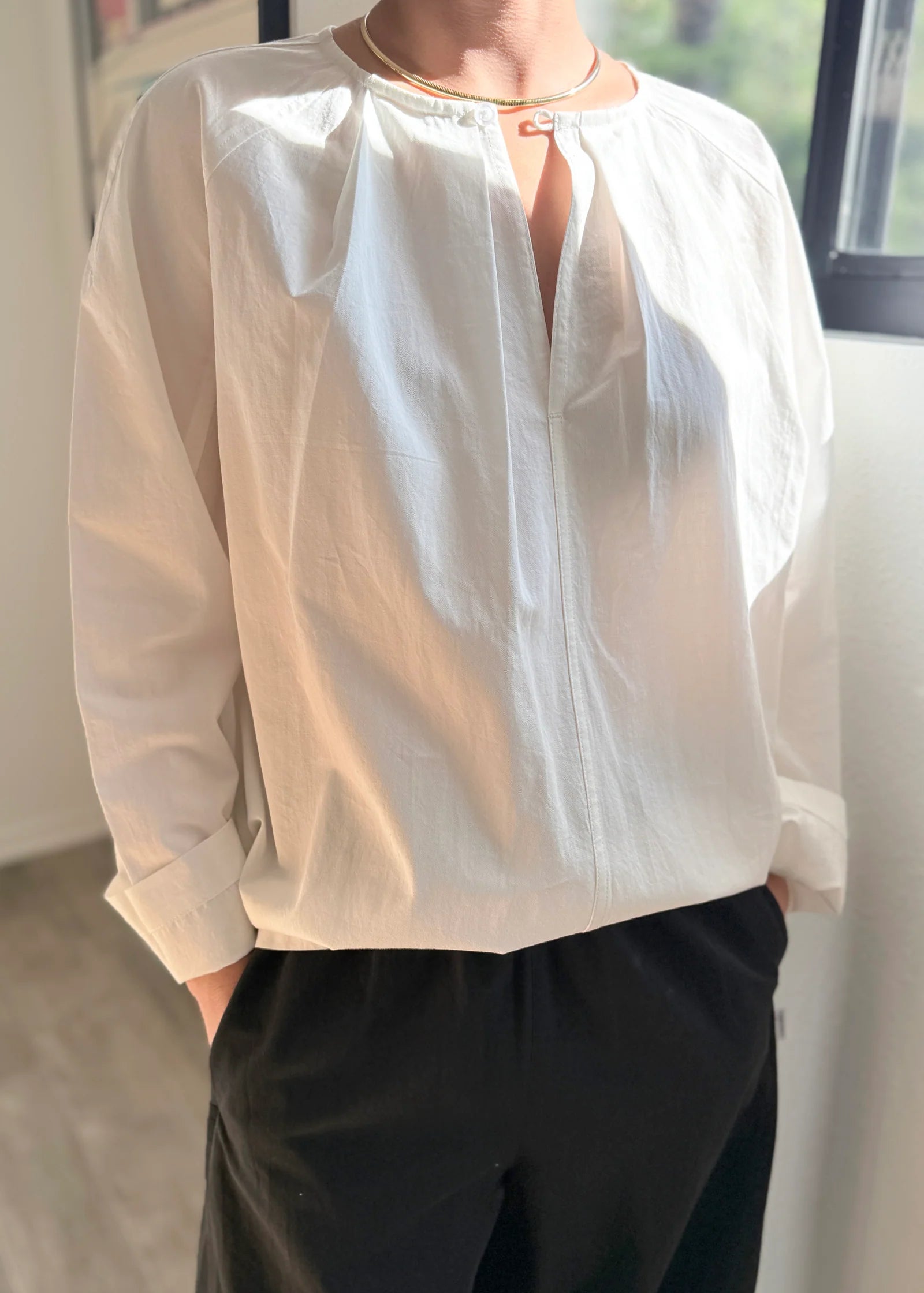 Person wearing a light-colored blouse with long sleeves and dark pants indoors.