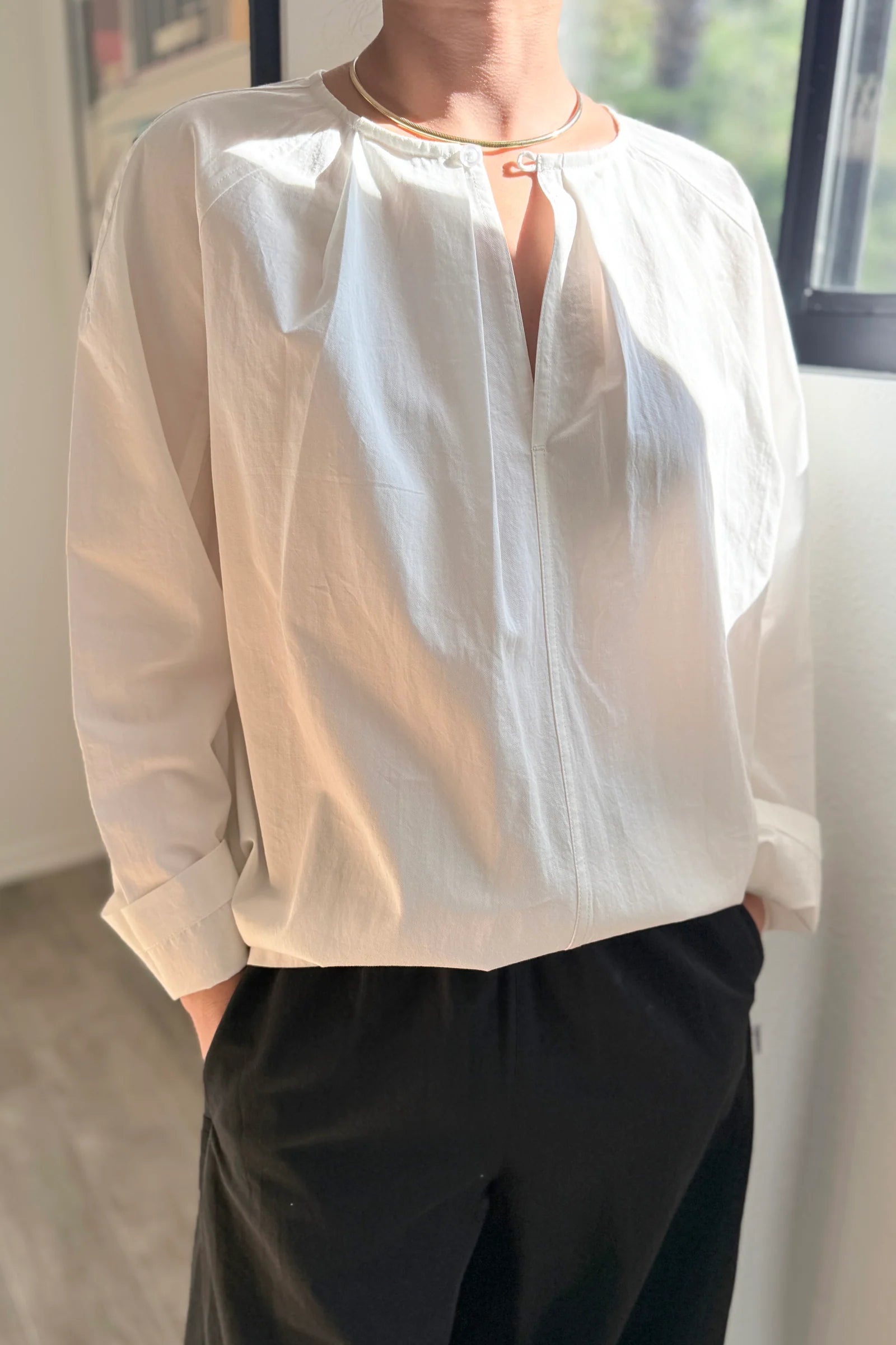 Person wearing a light-colored blouse with long sleeves and dark pants indoors.